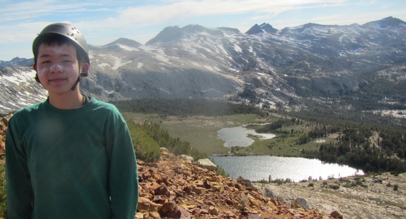 a person wearing a helmet and sunscreen poses for a photo in front a body of water and mountainous landscape